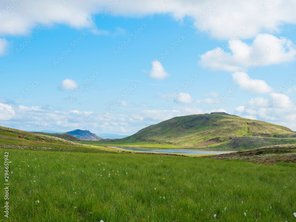 Fototapeta premium Blue lake, lush green grass meadow and hills at Reykjadalur hot springs area, South Iceland near Hveragerdi city. Summer sunny morning, blue sky.