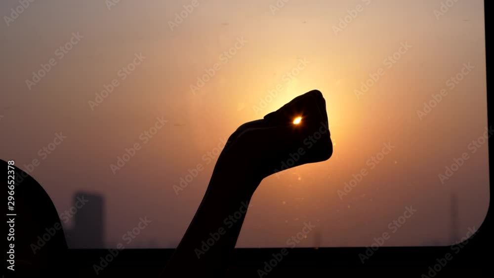 Passenger woman hold sun in hand, silhouetted shot of fist against window, sunset sky seen on background, halo of sunlight around arm. Tourist girl travel by high speed train at evening time