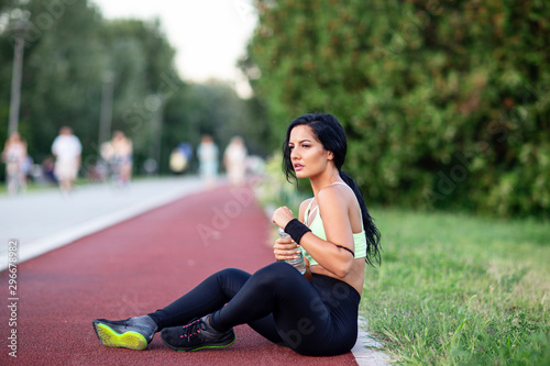 beautiful young girl sitting at the tartan track and relax after jogging with bottle of water