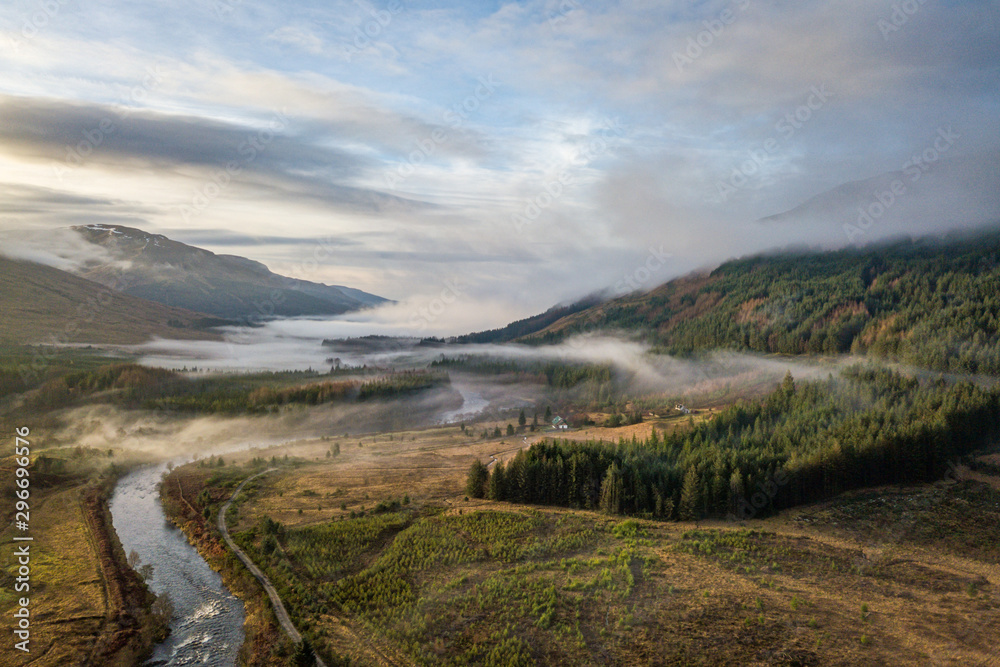Sun is setting as low hanging clouds run through the River Orchy, a pine forest and the hills in the distance on a partially cloudy day in the Highlands of Scotland.