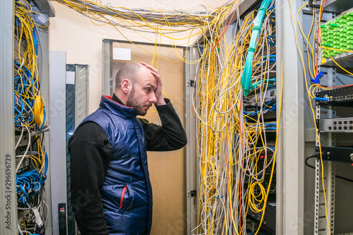 A sad man put his hand to his face at the sight of problems with laying wires in the server room. Incorrect connection of communication equipment in the data center. Wrong work photo