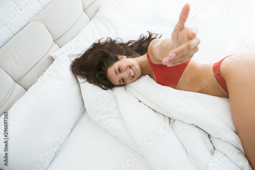 young beautiful woman in red sexy underwear lying in bed in bright bedroom smiling showing follow me sign