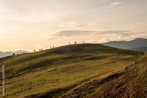 People watching the sun set on green grassy fields on Arthur’s Seat on a partially sunny day in Edinburgh, Scotland.