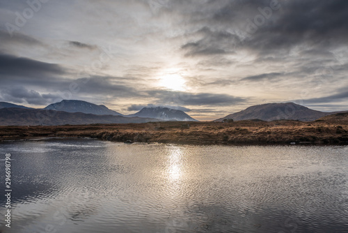 Wetlands and wintery grass fields as the sun starts to set over the Black Mount mountain range on a partially cloudy day in the Scottish Highlands.