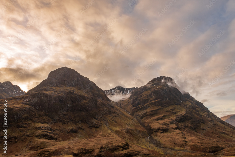Two of the three mountain peaks called The Three Sisters, covered in brown wintery vegetation on a partially cloudy day in Scottish Highlands.