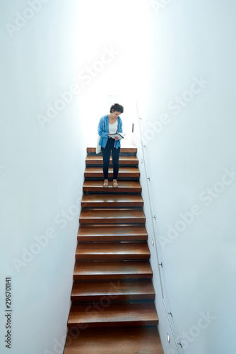 Young woman going down some stairs while reading some documents, the stairs are very white and mysterious.