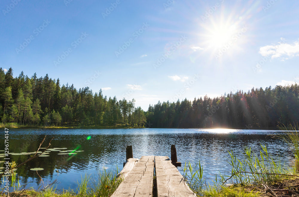 Fototapeta Traditional Finnish and Scandinavian view. Beautiful lake on a summer day and an old rustic wooden dock or pier in Finland. Sun shining on forest and woods in blue sky.
