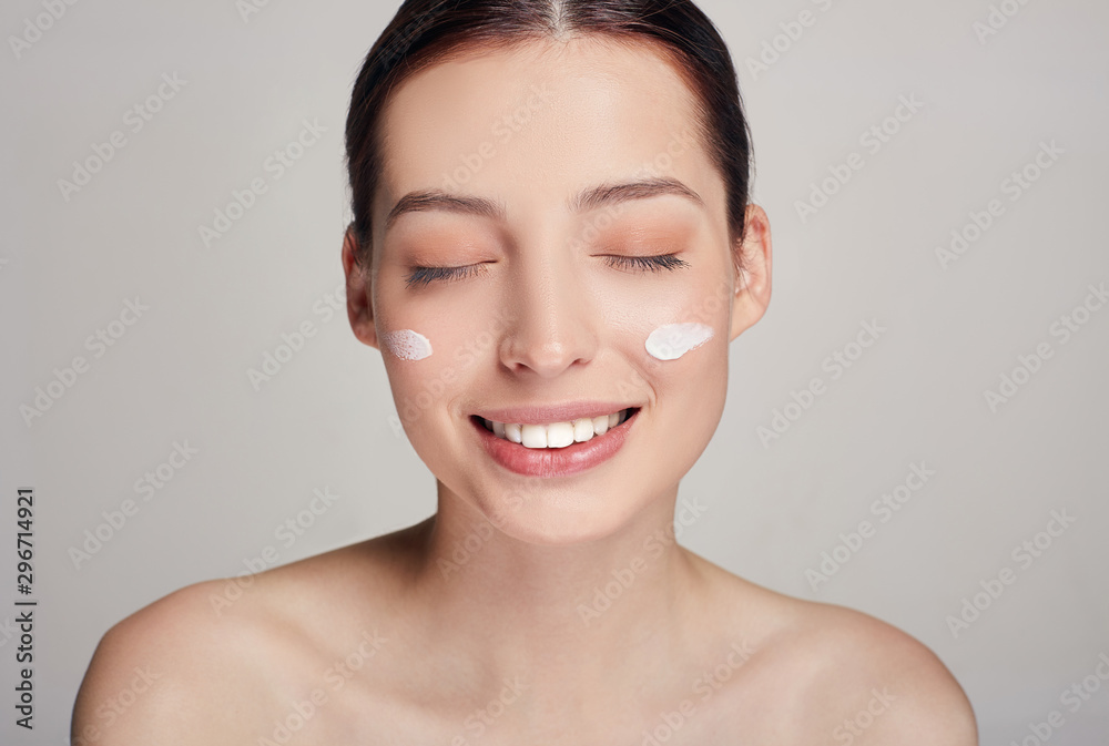 Smiling young beautiful girl with brown hair and clean skin with closed eyes and cream on her cheeks. Expressive facial expressions. Smiling young beautiful girl with brown hair and clean skin with closed eyes and cream on her cheeks. Expressive facial expressions.