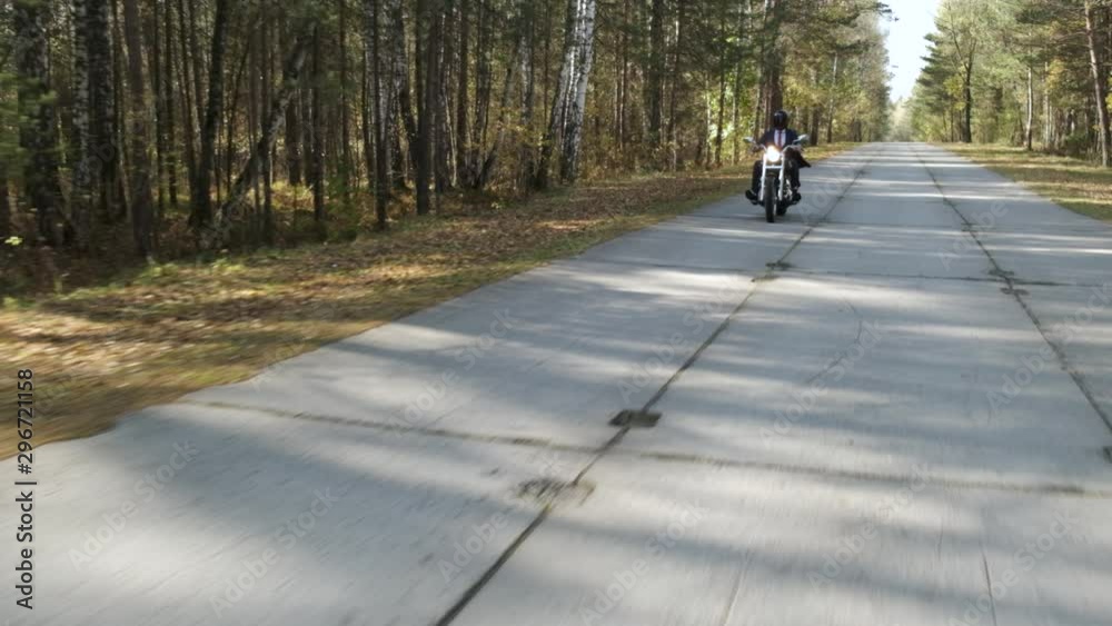 Cool motorcyclist in classic business suit is rides along autumn road, bright light hits between trees, lot of fallen leaves on side of road, man control chrome motorcycle in slow motion, overall plan Cool motorcyclist in classic business suit is rides along autumn road, bright light hits between trees, lot of fallen leaves on side of road, man control chrome motorcycle in slow motion, overall plan