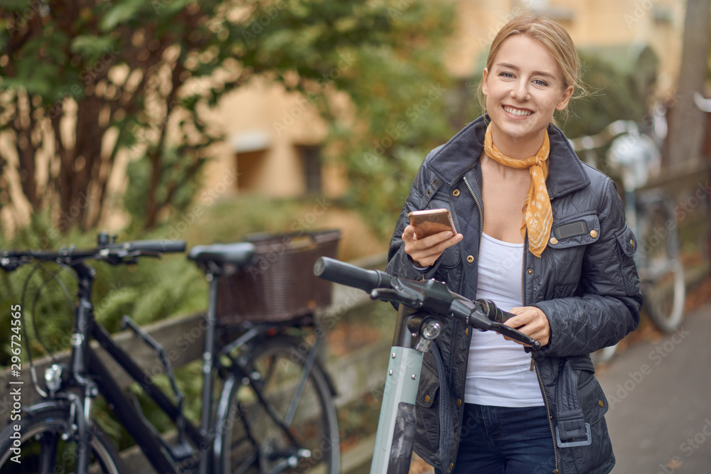 Fototapeta premium Happy young woman in casual grey jacket looking at camera and smiling, standing in the street with an e-scooter with mobile phone in her hands.