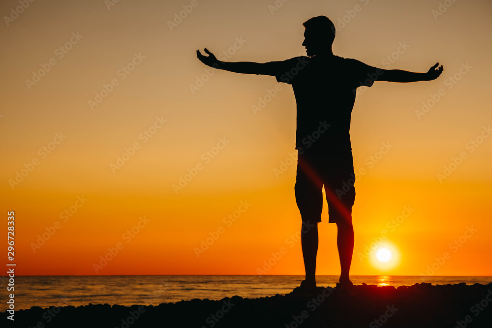 Silhouette of a man stands and contemplating on beach on sea background and sunset flinging his hands to the side. Shoot from the back. Concept of freedom relaxation. Place for text or advertising