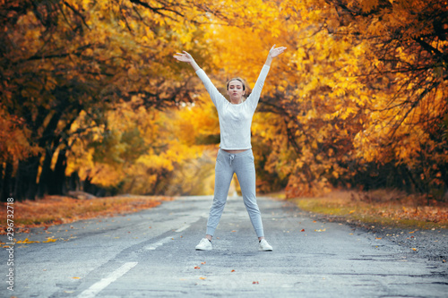 slim young woman in sportswear exercises and stretches hands up satanding on asphalt road  girl engaged in sport outdoors on background of autumn foliage  concept healthy lifestyle and female beauty