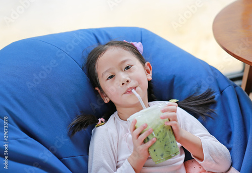 Adorable little Asian child girl drinking iced Matcha Green Tea lying on sofa at cafe. photo