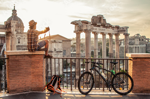 Canvas Print Young man tourist with backpack and bike sitting at Roman Forum at sunrise taking picture with vintage camera