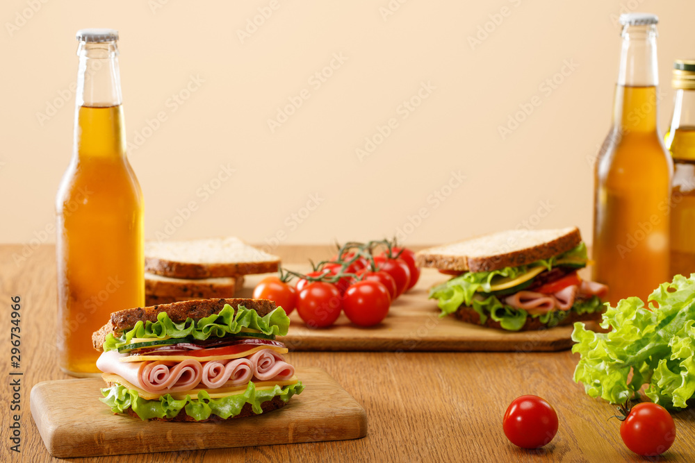 selective focus of fresh sandwich with lettuce, ham, cheese, bacon and tomato near bottles of beer at wooden table isolated on beige selective focus of fresh sandwich with lettuce, ham, cheese, bacon and tomato near bottles of beer at wooden table isolated on beige