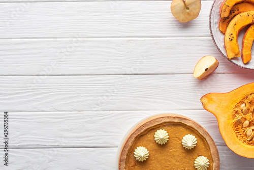delicious pumpkin pie with whipped cream near raw and baked pumpkins, whole and cut apples on white wooden table