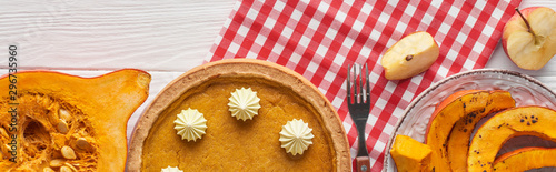 Fototapeta Naklejka Na Ścianę i Meble -  panoramic shot of pumpkin pie with whipped cream on plaid napkin near baked and raw pumpkins, cut apple and fork on white woode table