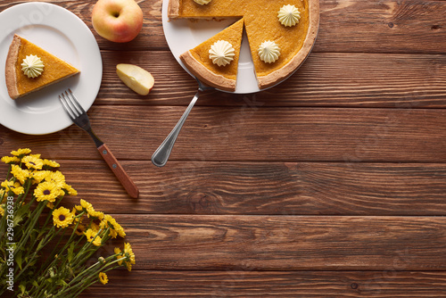 delicious pumpkin pie with whipped cream on plates with spatula and fork near cut and whole apples, and yellow flowers on brown wooden table