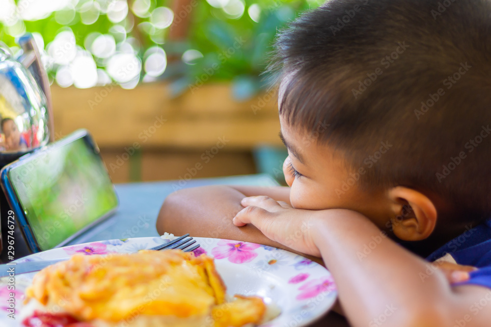 Soft focus. Dinner time at the restaurant, Asian little boy eating rice with omelette and playing mobile phone in the same time. Soft focus. Dinner time at the restaurant, Asian little boy eating rice with omelette and playing mobile phone in the same time.