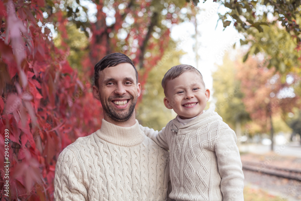 Dad and mvlenky son walk in the autumn park. Bright, warm autumn. Red leaves. Cozy. Portrait of dad and son in knitted sweaters.