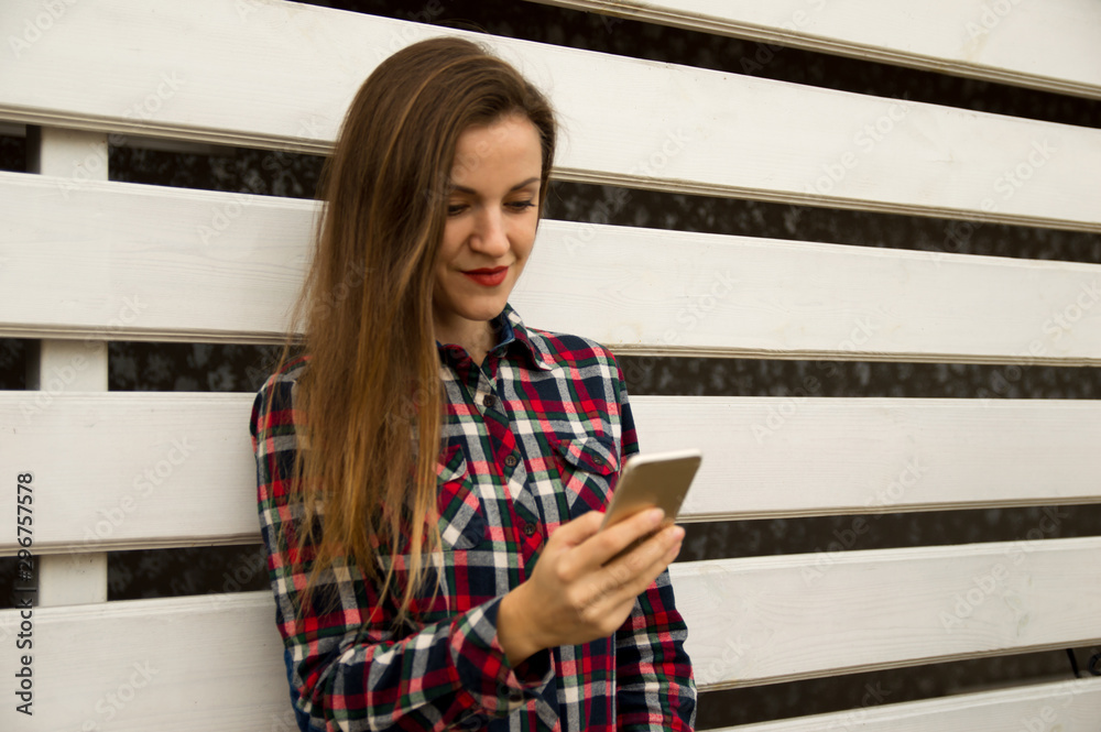 Young beautiful girl stands near a white wall, smiles and holds a mobile phone, looks at the phone screen.
