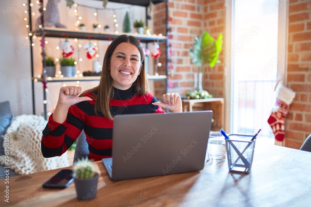 Beautiful woman sitting at the table working with laptop at home around christmas lights looking confident with smile on face, pointing oneself with fingers proud and happy. Beautiful woman sitting at the table working with laptop at home around christmas lights looking confident with smile on face, pointing oneself with fingers proud and happy.