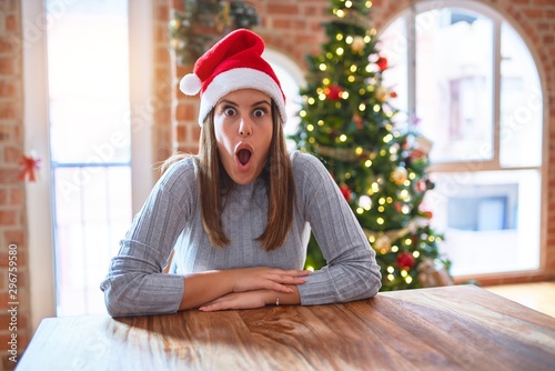 Young beautiful woman wearing santa claus hat at the table at home around christmas decoration afraid and shocked with surprise expression, fear and excited face.