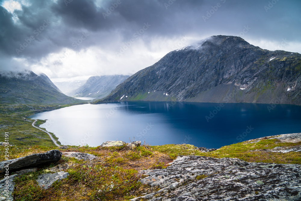 View of lake between Norwegian fjords and mountains with a green grass, blue water and cloudy sky