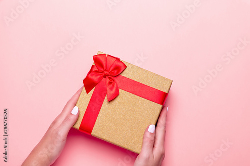 Woman manicured hands holding red and golden wrapped present or giftbox on pastel pink background, copy space, top view, flat lay. Background for Valentine's Day, Mother's Day.