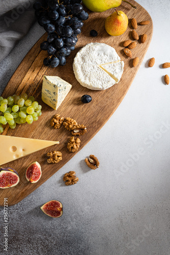 Cheese plate: Maasdam, Camembert cheese, blue cheese, figs, walnuts, almonds, pears and grapes on wooden board on gray background. Copy space for text. Top view, falt lay. Appetizer, menu photo