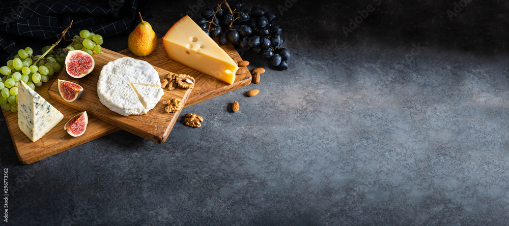 Cheese plate served with camembert, brie, blue cheese, maasdam, grapes, pear, figs and nuts on a wooden board on gray background. Side view, copy space. Banner - obrazy, fototapety, plakaty 