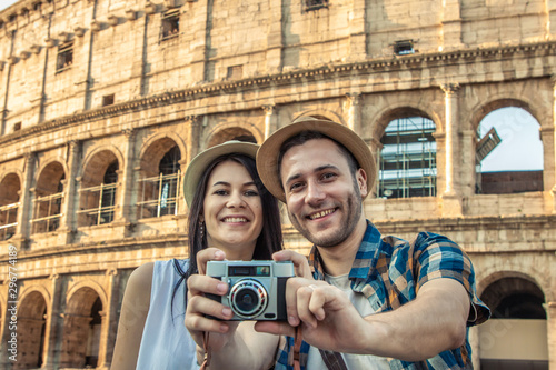 Lovely couple of tourist sitting on a wall kissing at Colosseum while taking pictures looking at camera. Rome, Italy