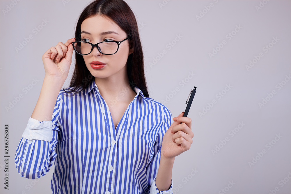 In the background is a pleasant young business woman with brown hair and glasses. girl holds a black pen in her hand. other hand touches her glasses. In the background is a pleasant young business woman with brown hair and glasses. girl holds a black pen in her hand. other hand touches her glasses.
