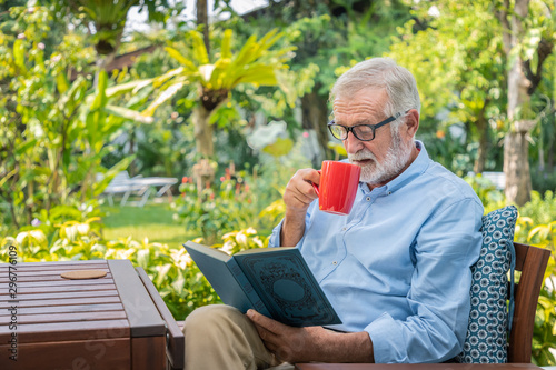 Senior elderly man reading book drinking mug of coffee in garden