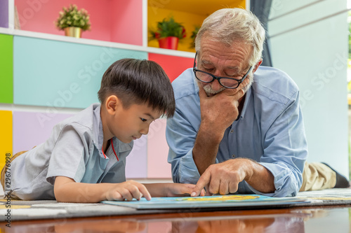 Happy boy grandson reading book with old senior man grandfather at home