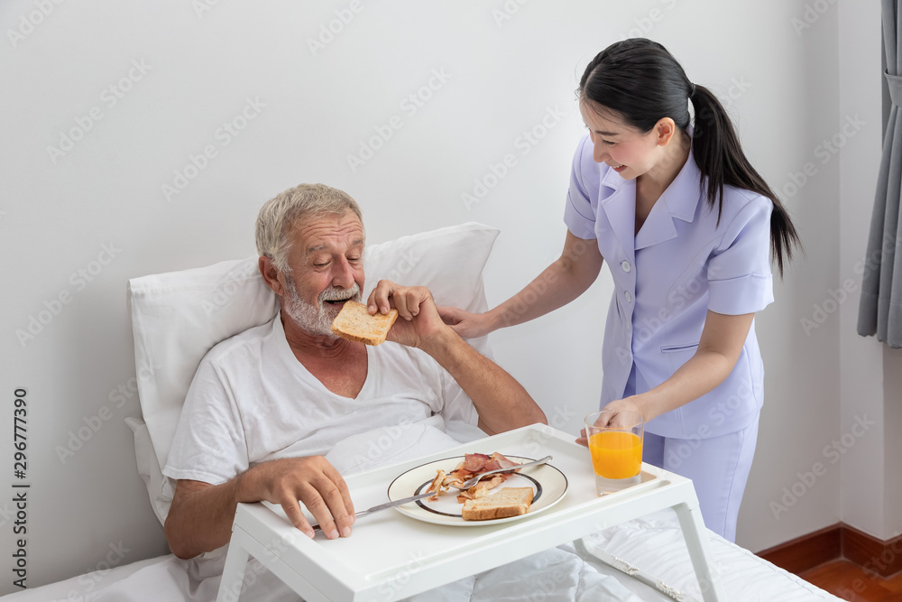Happy nurse assist and help elderly senior man to eat breakfast on bed in bedroom at nursing home Happy nurse assist and help elderly senior man to eat breakfast on bed in bedroom at nursing home