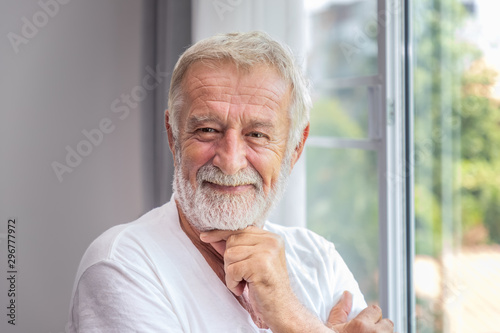 Senior elderly man standing at window in bedroom after waking up in morning, looking camera photo