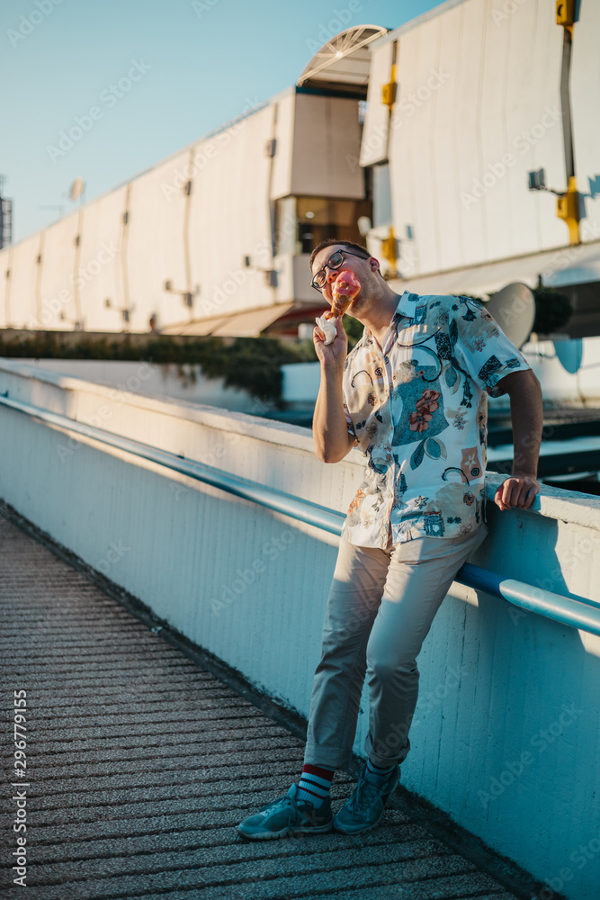 Young fashionable man eating ice cream in a cone in the city at sunset - obrazy, fototapety, plakaty 