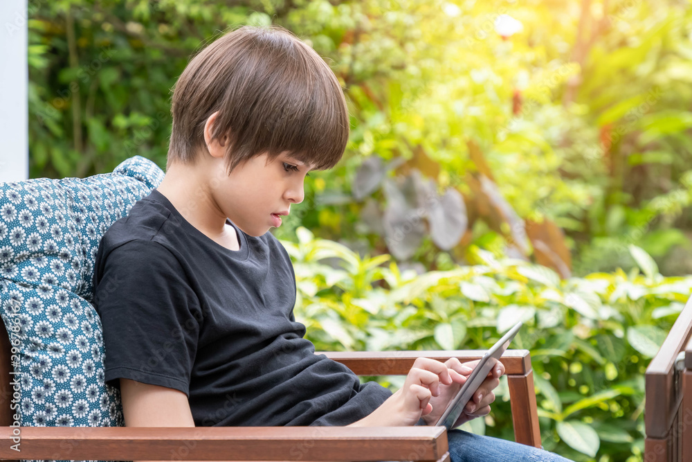 Young cute Caucasian boy using digital tablet sitting on couch in garden at home Young cute Caucasian boy using digital tablet sitting on couch in garden at home