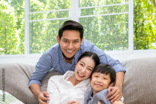 Happy Asian father, mother, and son sitting together on sofa in living room, looking at camera; dad hugging mom and boy; family relationship concept