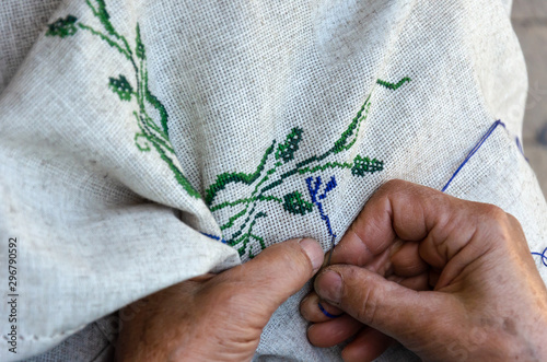 Hands of an elderly woman embroidering a cross-stitch floral pattern on linen fabric. Embroidery, handwork, needlecraft concept. Closeup