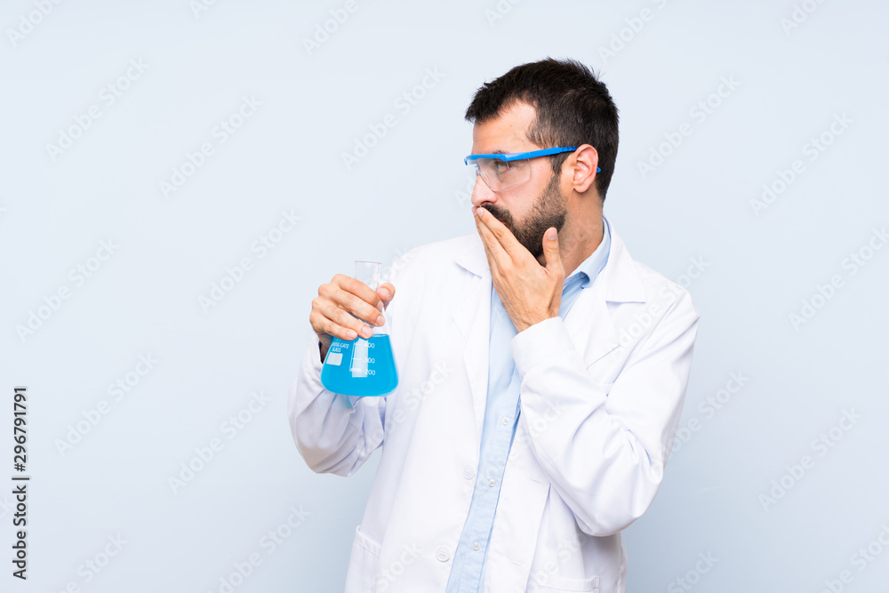 Young scientific holding laboratory flask over isolated background covering mouth with hands Young scientific holding laboratory flask over isolated background covering mouth with hands