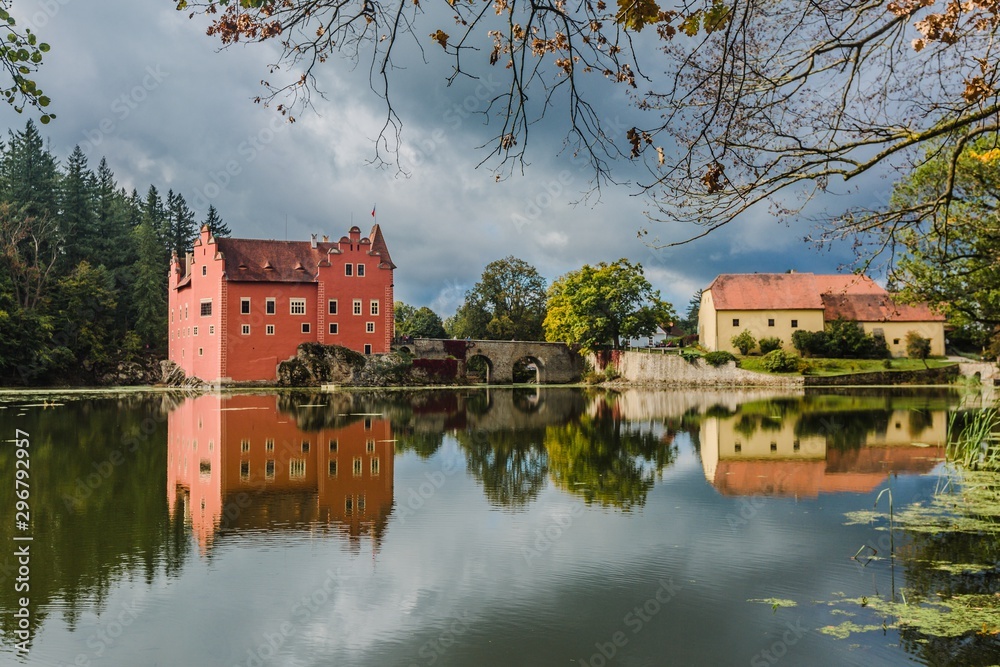 Cervena Lhota, Czech Republic - September 28 2019: View of famous red castle standing on a rock in the middle of a lake. Sunny day and blue sky with clouds. Reflection of buildings in water.
