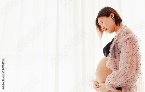 Happy pregnant woman standing near window and touching her belly at home