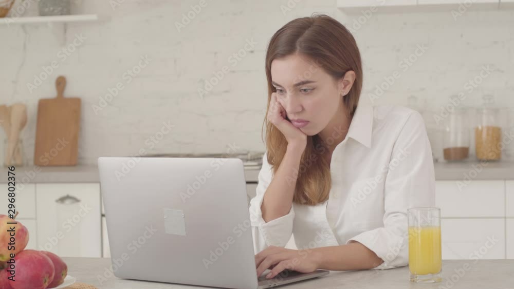 Young tired Caucasan businesswoman sitting at the kitchen with laptop and sighing. Young pretty girl in white shirt working online from home. Young tired Caucasan businesswoman sitting at the kitchen with laptop and sighing. Young pretty girl in white shirt working online from home.