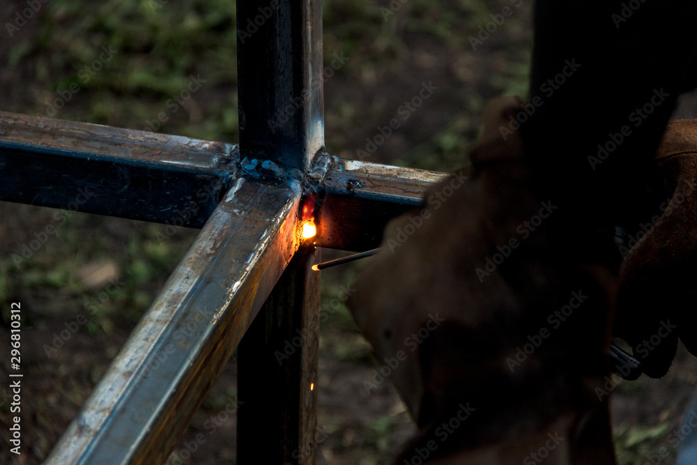 Naklejka premium A strong man is a welder, welding mask and welders leathers, a metal product is welded with a arc welding machine at the construction site, blue sparks fly to the sides