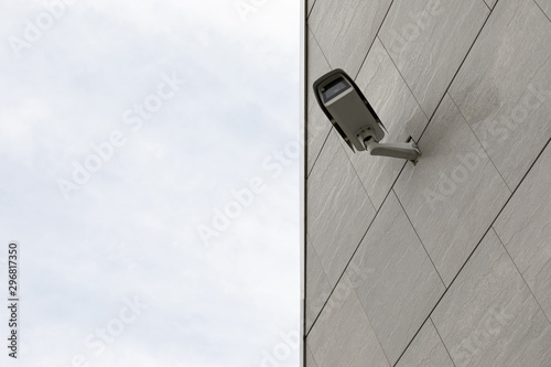 Safety cctv camera on gray tiled building facade, sky on other half of photo, diagonal view.