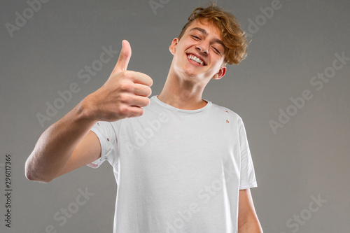 handsome emotional teenager boy posing in studio against gray, guy in a gray t-shirt shows finger like