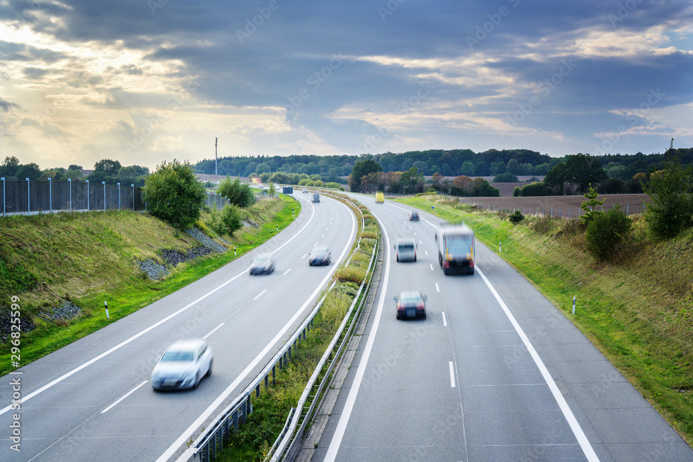 Fototapeta premium Country highway with driving cars and trucks in motion blur, concept for traffic, transport and environmental protection, copy space in the sky with clouds