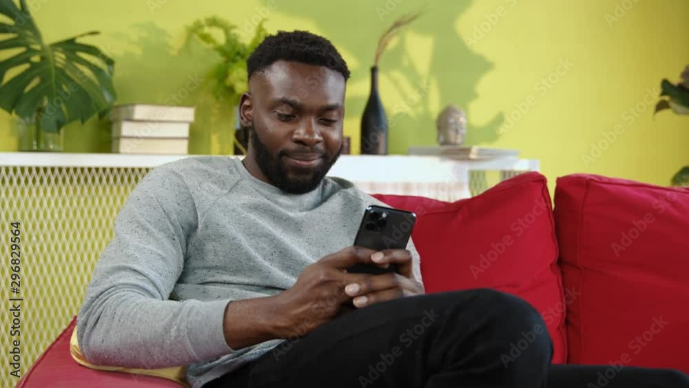 Cheerful young afro-american man browsing content in modern smartphone sitting on red sofa in stylish apartment living room. Cheerful young afro-american man browsing content in modern smartphone sitting on red sofa in stylish apartment living room.